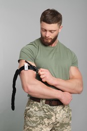 Photo of Soldier in military uniform applying medical tourniquet on arm against light grey background
