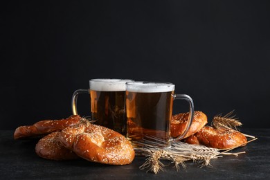 Photo of Tasty pretzels, glasses of beer and wheat spikes on black table against dark background, space for text