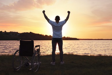 Photo of Man raising hands up to sky near wheelchair at sunset, back view. Healing miracle
