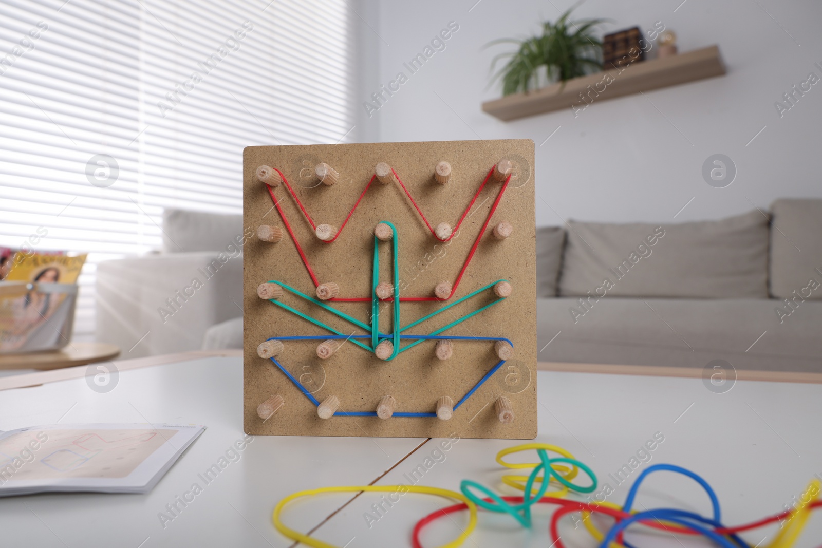 Photo of Wooden geoboard with flower made of rubber bands on white table in room. Motor skills development