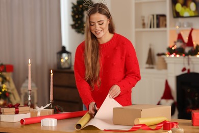 Beautiful young woman decorating Christmas gift with wrapping paper at table in room