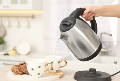 Photo of Woman pouring hot water from electric kettle into cup in kitchen, closeup. Space for text