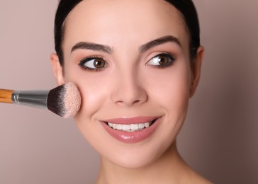 Photo of Beautiful young woman applying face powder with brush on dusty rose background, closeup
