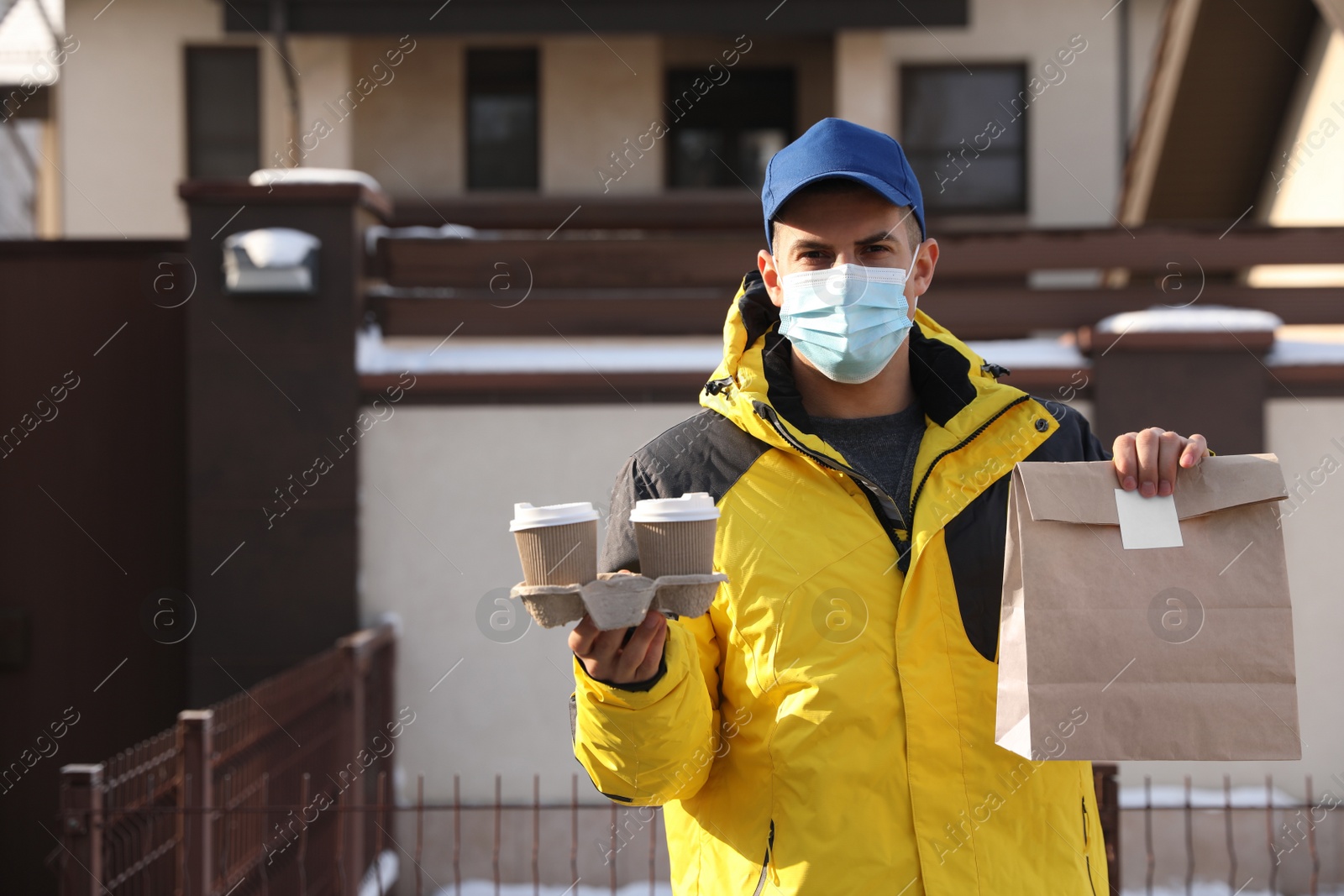 Photo of Courier in medical mask holding takeaway food and drinks near house outdoors. Delivery service during quarantine due to Covid-19 outbreak