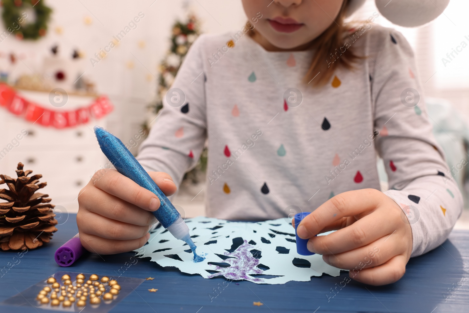 Photo of Little child making Christmas craft at blue wooden table in decorated room, closeup
