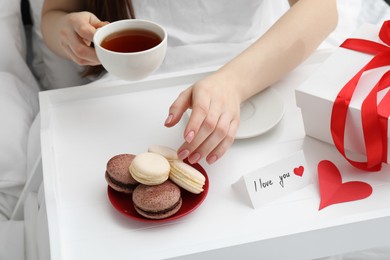 Photo of Tasty breakfast served in bed. Woman with tea, macarons, gift box and I Love You card at home, closeup