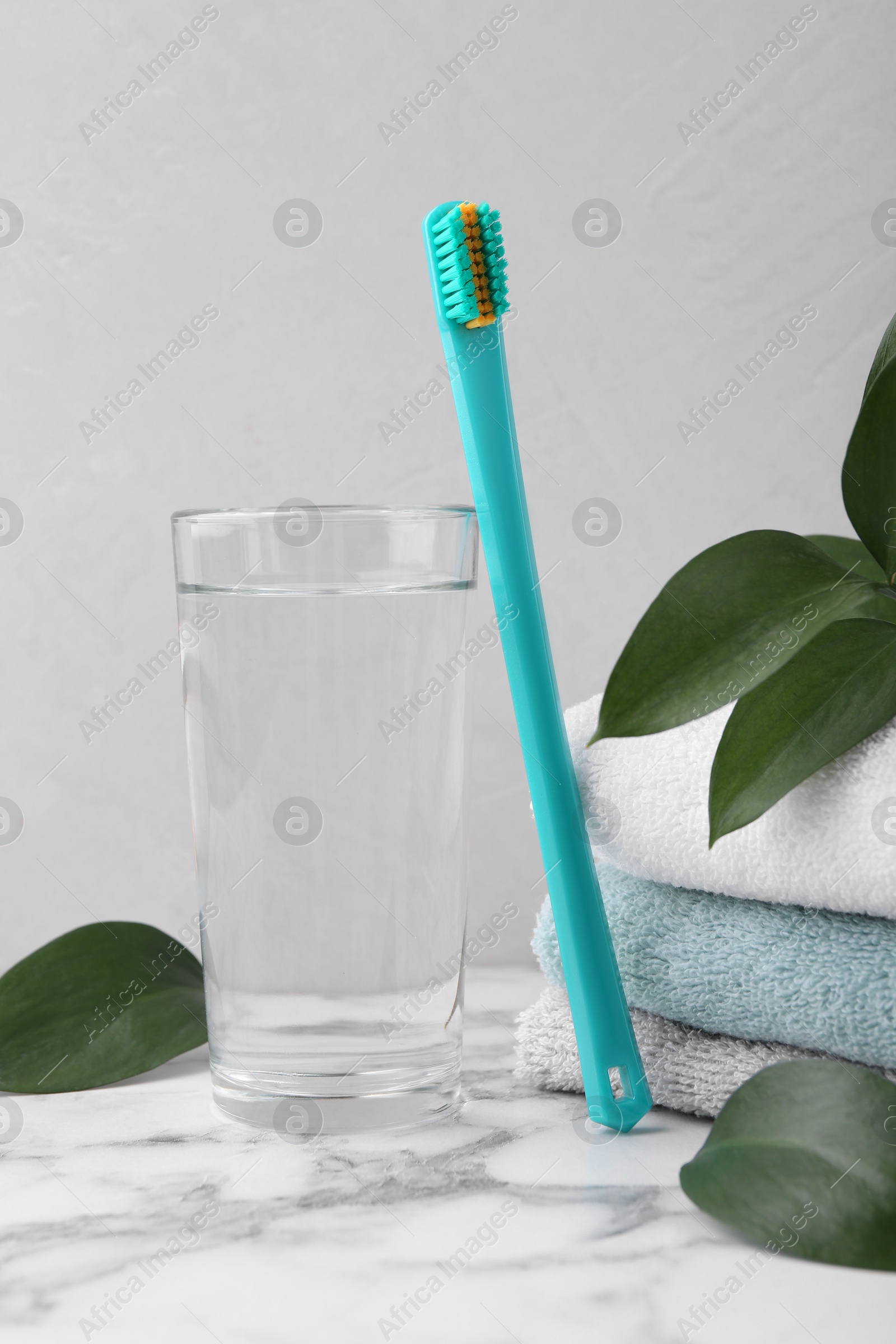 Photo of Plastic toothbrush, glass of water, towels and green leaves on white marble table
