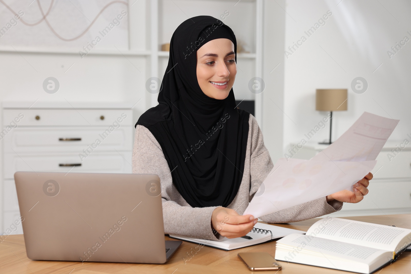 Photo of Muslim woman working near laptop at wooden table in room