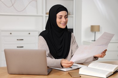 Photo of Muslim woman working near laptop at wooden table in room