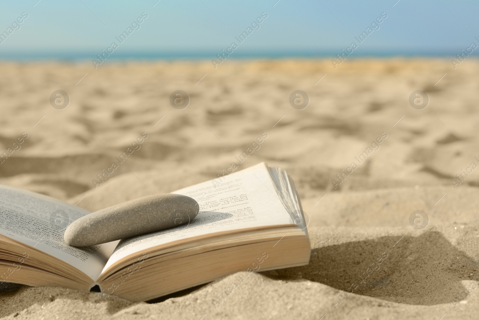 Photo of Open book with stone on sandy beach near sea, closeup. Space for text