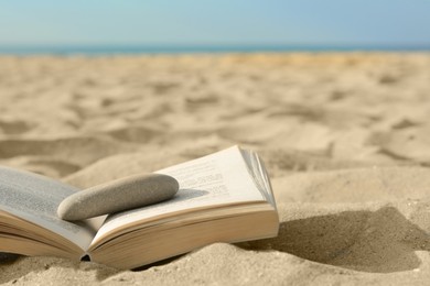 Open book with stone on sandy beach near sea, closeup. Space for text