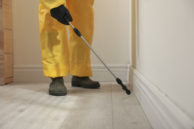 Photo of Pest control worker in protective suit spraying insecticide on floor indoors, closeup