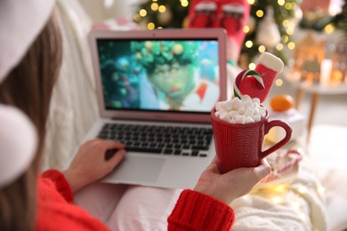 MYKOLAIV, UKRAINE - DECEMBER 25, 2020: Woman with sweet drink watching The Grinch movie on laptop at home, closeup. Cozy winter holidays atmosphere