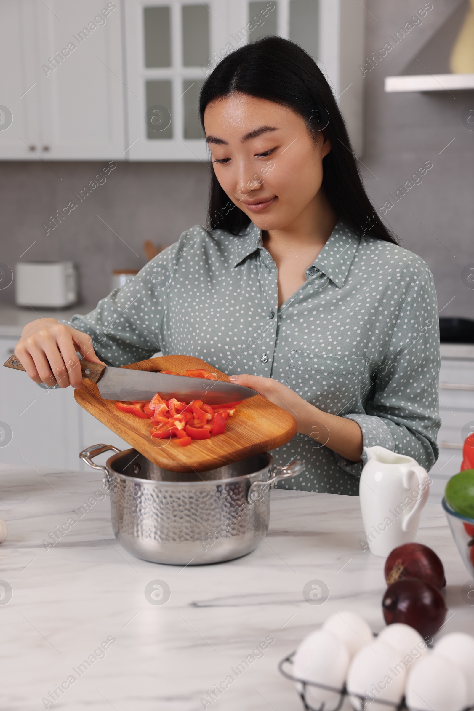 Photo of Cooking process. Beautiful woman adding cut bell pepper into pot at white marble table in kitchen