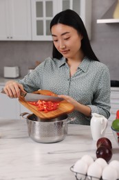 Photo of Cooking process. Beautiful woman adding cut bell pepper into pot at white marble table in kitchen