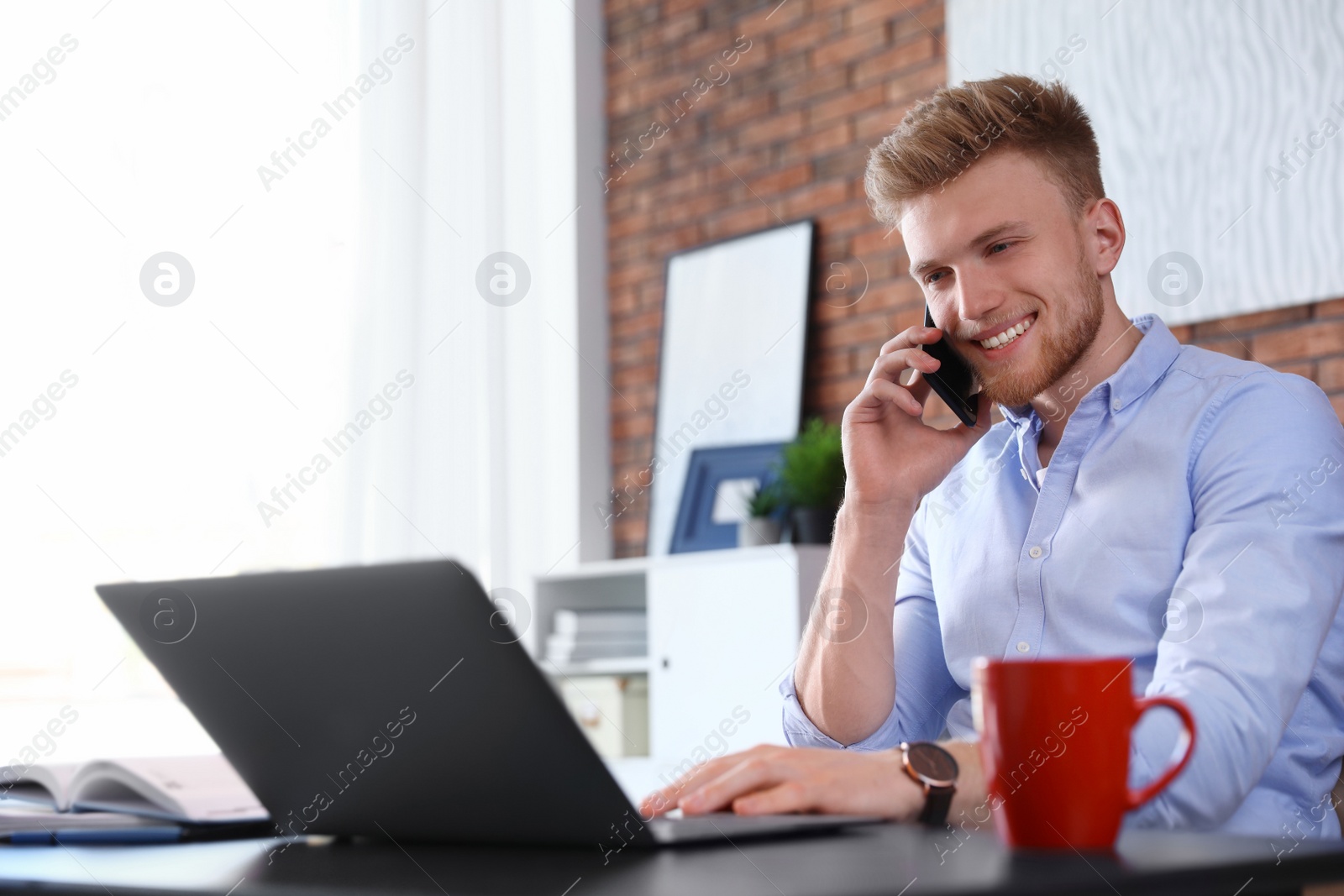 Photo of Young man talking on phone while using laptop at table indoors