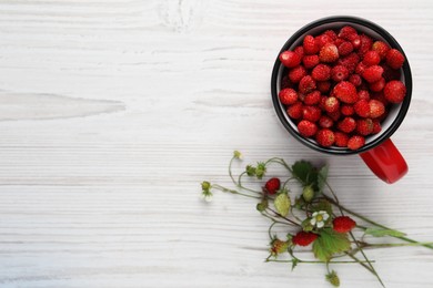 Photo of Fresh wild strawberries in mug near stems with flowers and leaves on white wooden table, flat lay. Space for text