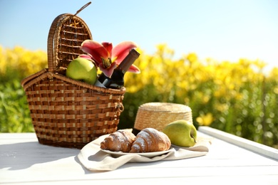 Photo of Picnic basket with bottle of wine and food on white wooden table in lily field