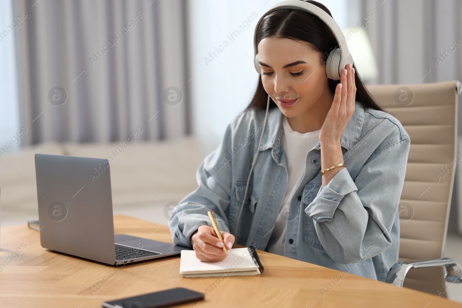 Photo of Young woman in headphones writing down notes during webinar at table in room