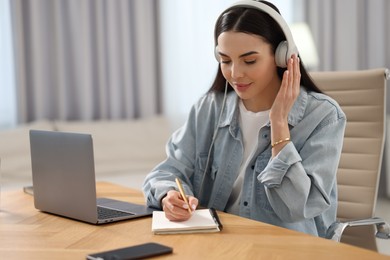 Photo of Young woman in headphones writing down notes during webinar at table in room