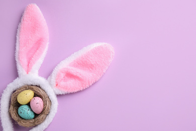 Photo of Headband with Easter bunny ears and dyed eggs in nest on violet background, flat lay. Space for text