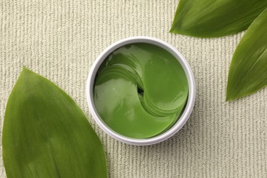 Photo of Jar of under eye patches and green leaves on light cloth, top view. Cosmetic product