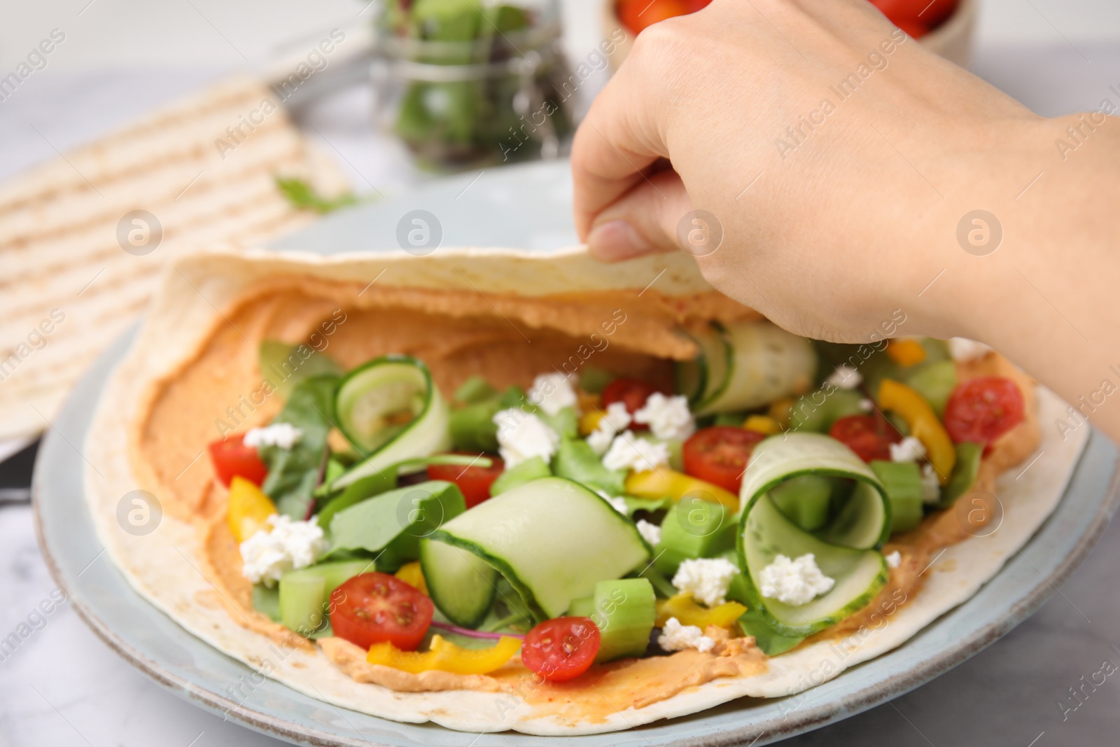 Photo of Woman making delicious hummus wrap with vegetables at table, closeup