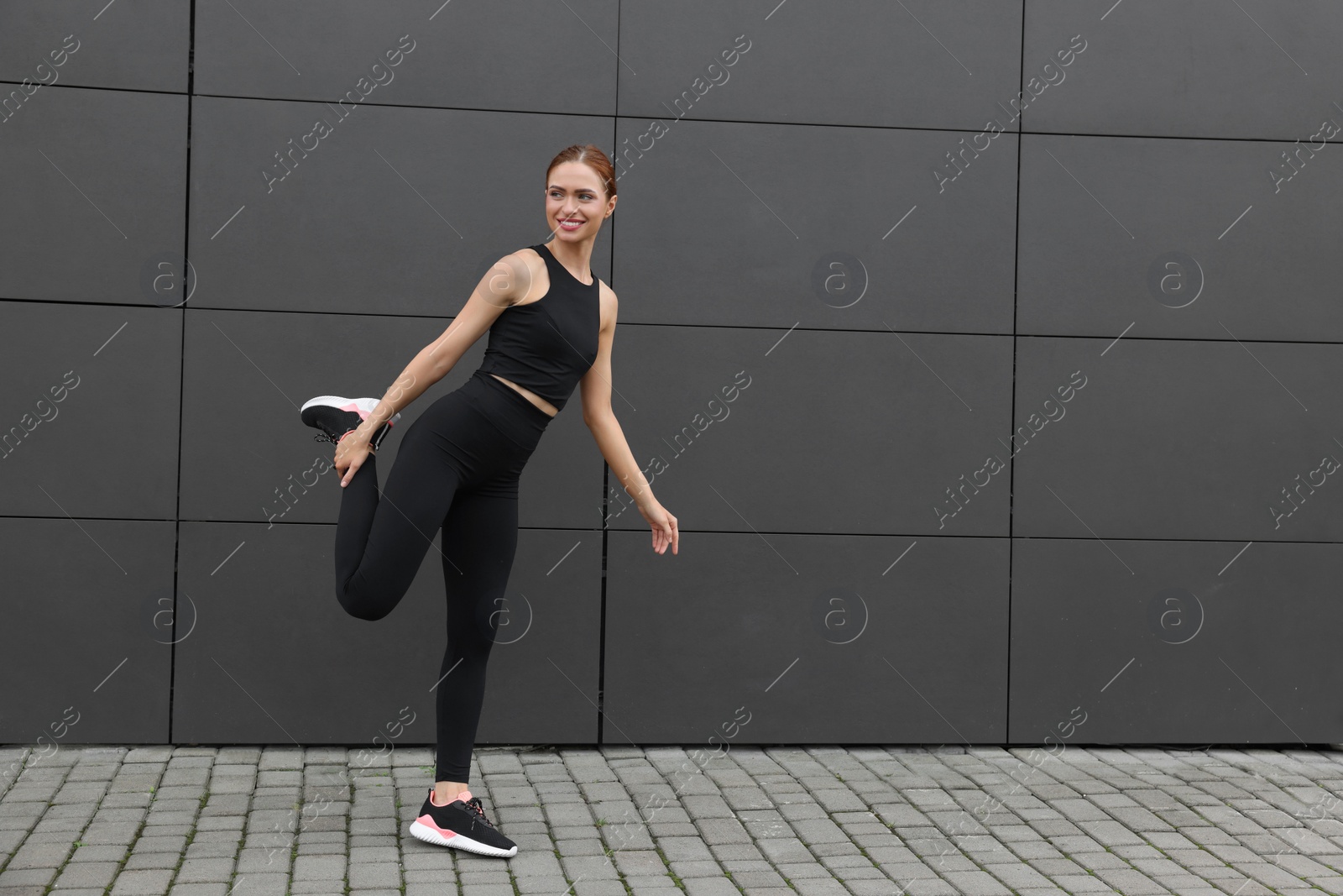 Photo of Beautiful woman in gym clothes doing exercises near dark grey wall on street, space for text
