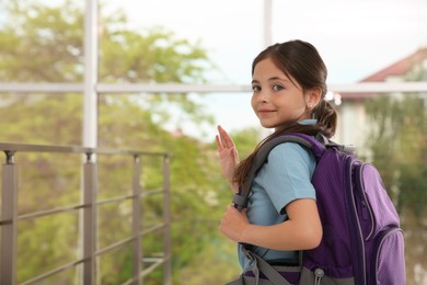 Photo of Happy little girl with schoolbag waving indoors. Space for text
