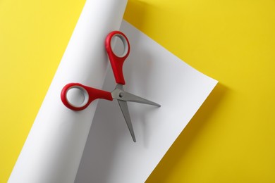 Red scissors and white paper on yellow background, top view