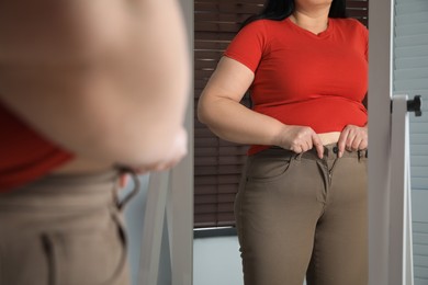 Photo of Overweight woman trying to button up tight trousers in front of mirror at home, closeup
