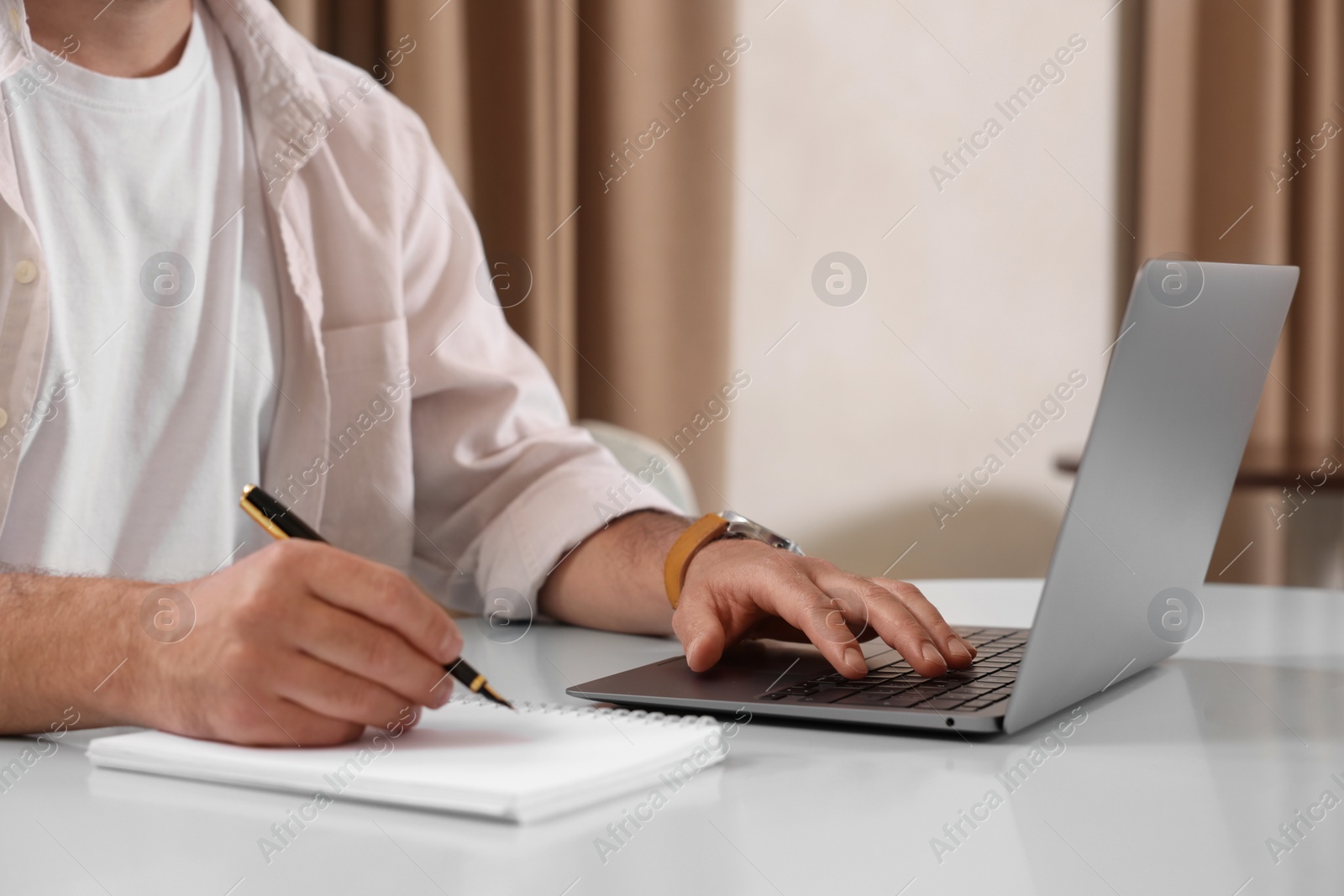 Photo of Man using laptop and writing something in notebook at table indoors, closeup