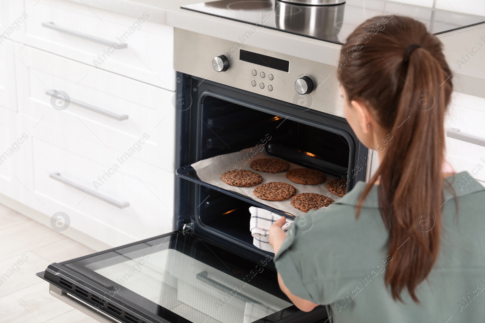 Photo of Young woman taking baking sheet with cookies from oven at home