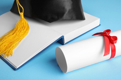 Photo of Graduation hat, open book and student's diploma on light blue background, closeup