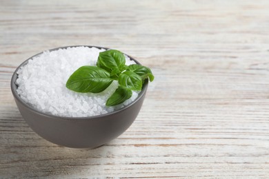 Photo of Bowl of natural sea salt and basil leaf on white wooden table, space for text