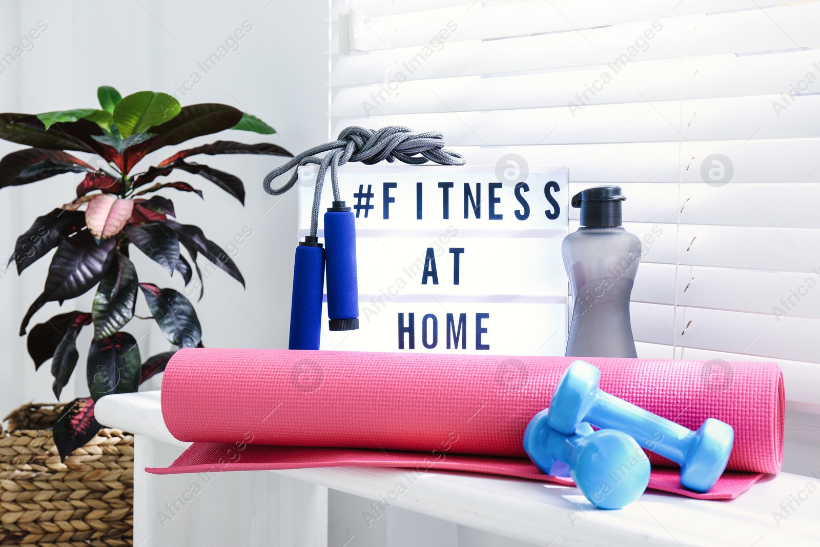 Photo of Sport equipment and lightbox with hashtag FITNESS AT HOME on window sill indoors. Message to promote self-isolation during COVID‑19 pandemic