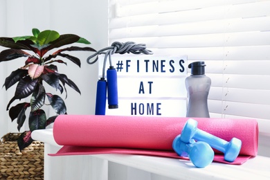 Sport equipment and lightbox with hashtag FITNESS AT HOME on window sill indoors. Message to promote self-isolation during COVID‑19 pandemic