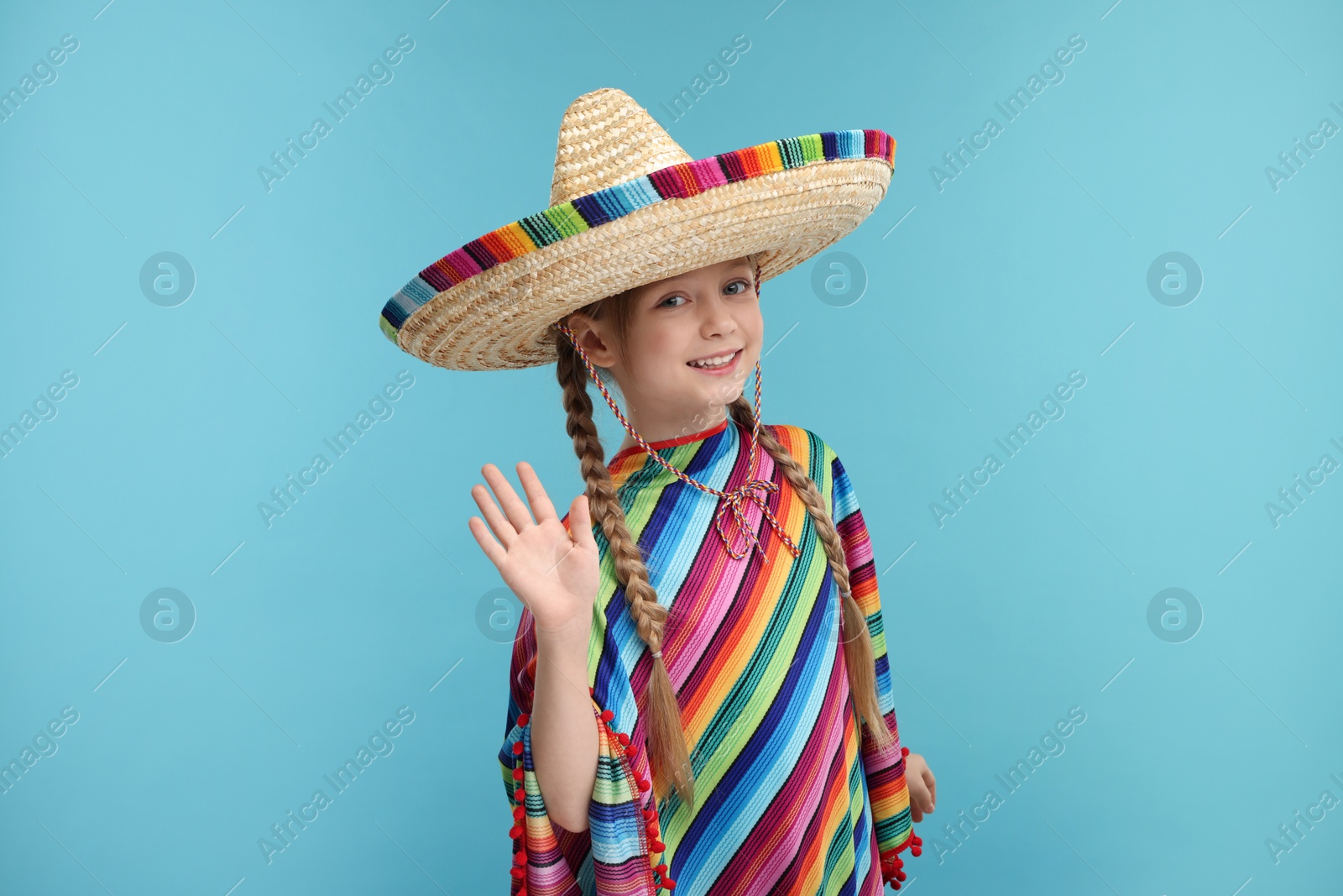 Photo of Cute girl in Mexican sombrero hat and poncho waving hello on light blue background