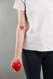 Blood donation concept. Woman with adhesive plaster on arm holding red heart against grey background, closeup