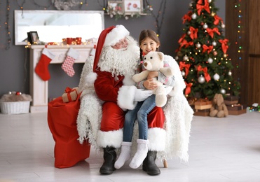 Photo of Little girl with teddy bear sitting on authentic Santa Claus' lap indoors