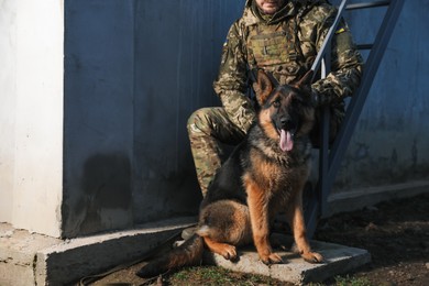 Photo of Ukrainian soldier with German shepherd dog sitting outdoors, closeup. Space for text