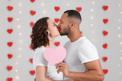Photo of Lovely couple with pink paper heart kissing indoors. Valentine's day celebration
