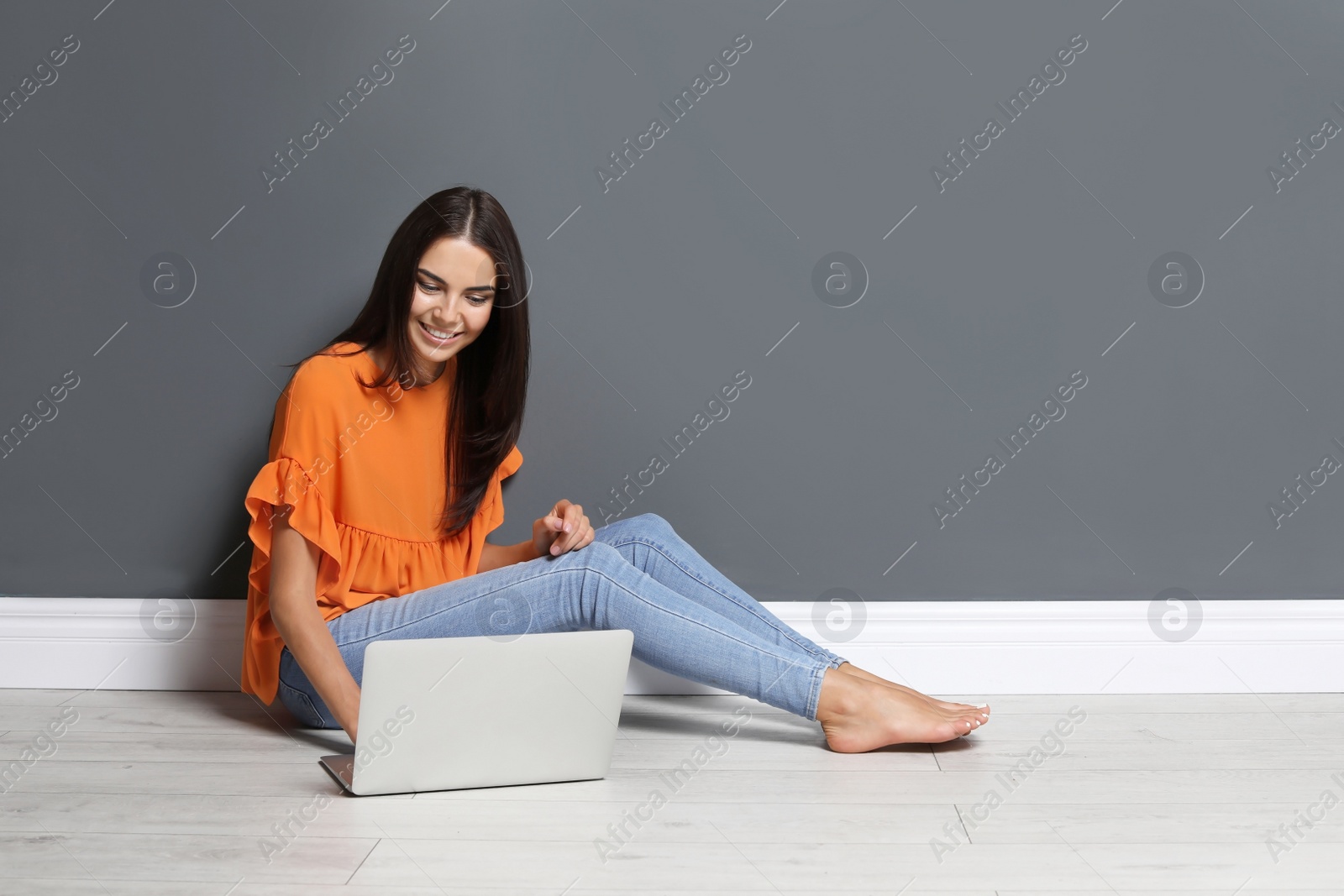 Photo of Young woman with modern laptop sitting on floor near grey wall
