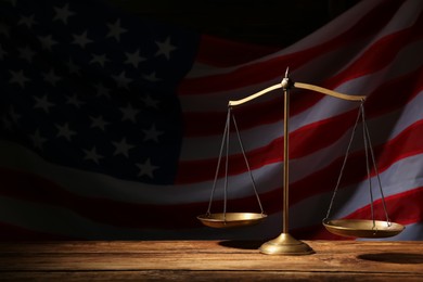Photo of Scales of justice on wooden table against American flag in darkness, space for text