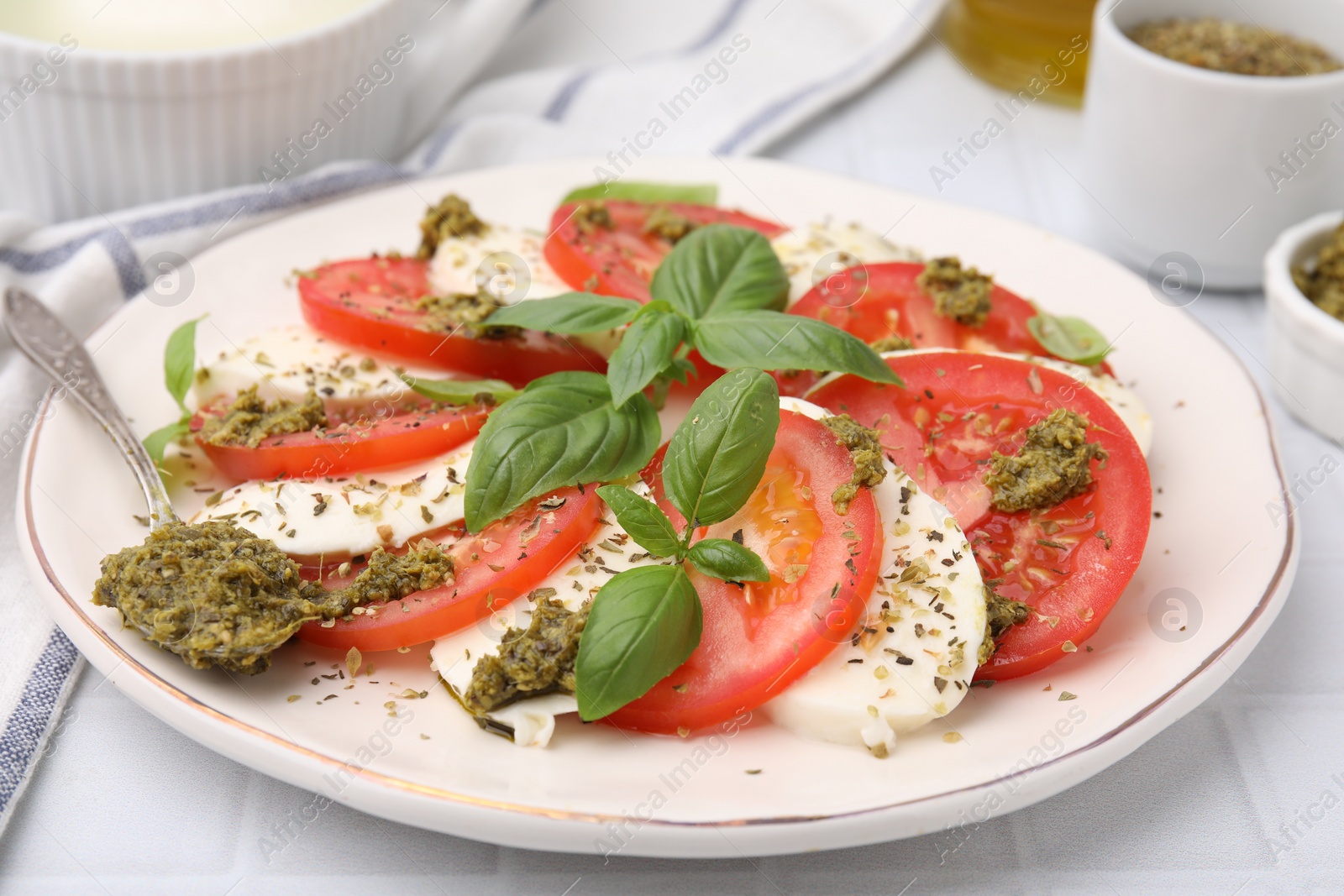 Photo of Plate of delicious Caprese salad with pesto sauce on white tiled table, closeup