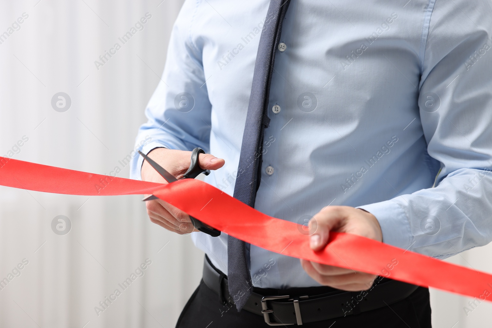 Photo of Man cutting red ribbon with scissors indoors, closeup