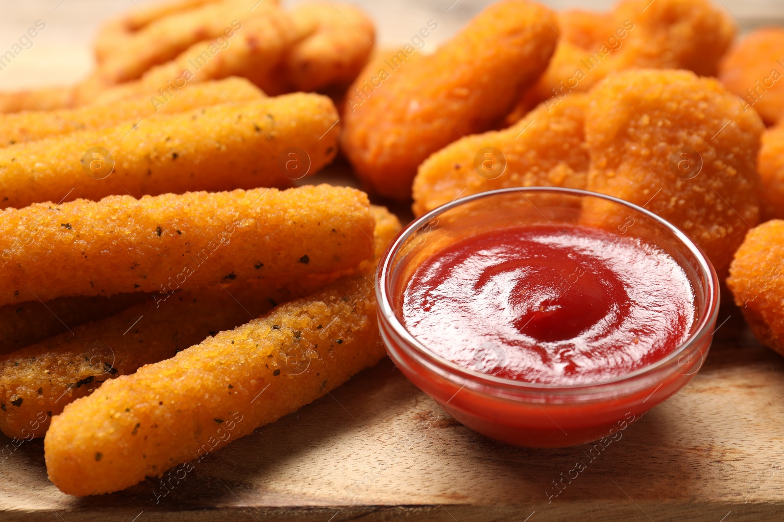 Photo of Bowl with tasty ketchup, cheese sticks and chicken nuggets on wooden table, closeup