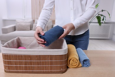 Photo of Woman putting rolled shirt into basket at table in room, closeup. Organizing clothes