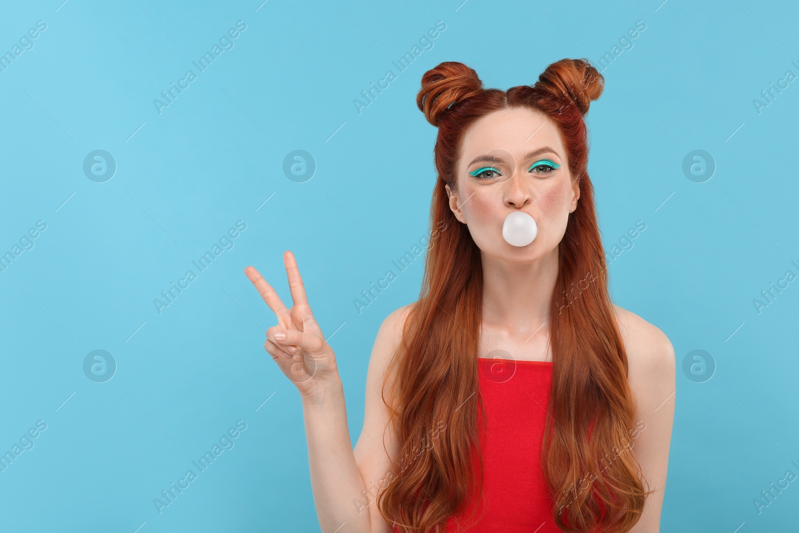 Photo of Portrait of beautiful woman with bright makeup blowing bubble gum and showing peace gesture on light blue background. Space for text