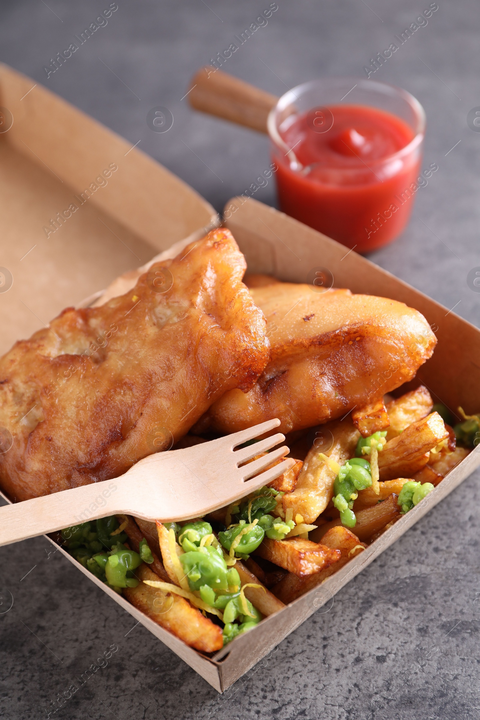 Photo of Tasty fish, chips, sauce and peas in paper box on grey table, closeup